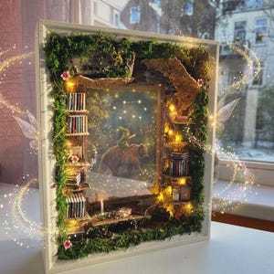 Może przedstawiać: Biała ramka typu shadow box przedstawiająca miniaturową bibliotekę z półkami pełnymi malutkich książek, zielonym mchem i akcentami z kory. W centrum znajduje się obraz postaci jadącej na niedźwiedziu, oświetlony ciepłymi światełkami LED.