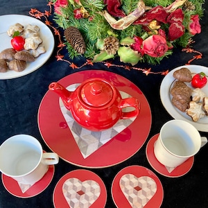 May include: Red and brown heart-shaped coasters with a geometric pattern. The coasters are on a black surface with a red teapot and a white mug.