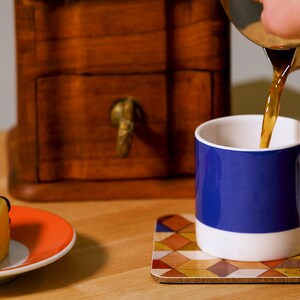 May include: A white mug with a blue band is being filled with coffee. A small yellow cake with chocolate stripes sits on an orange plate. A wooden box and a patterned coaster are also visible.
