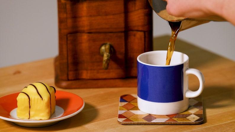 May include: A white mug with a blue band is being filled with coffee. A small yellow cake with chocolate stripes sits on an orange plate. A wooden box and a patterned coaster are also visible.