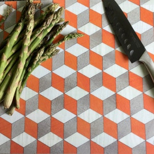 Könnte beinhalten: Nahaufnahme eines Schneidebretts mit einem geometrischen Muster aus orangefarbenen und grauen Quadraten. Ein Bund grüner Spargelspitzen und ein silbernes Messer liegen auf dem Brett.