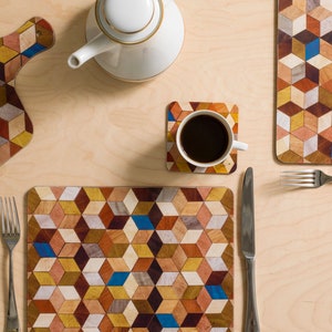 May include: Overhead shot of a wooden placemat, coaster, and cutting board with a geometric cube pattern in shades of brown, yellow, and blue. A white teapot and a cup of coffee sit on a light wood surface, with a fork and knife.