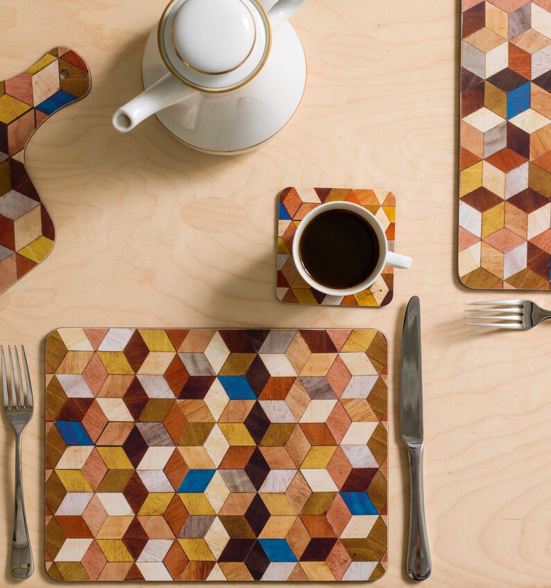 May include: Overhead shot of a wooden placemat, coaster, and cutting board with a geometric cube pattern in shades of brown, yellow, and blue. A white teapot and a cup of coffee sit on a light wood surface, with a fork and knife.