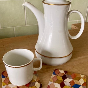 May include: A white ceramic coffee pot with a gold rim and lid, a matching cup, and two square coasters with a geometric pattern. The coasters feature a colorful mosaic design with various wood tones.
