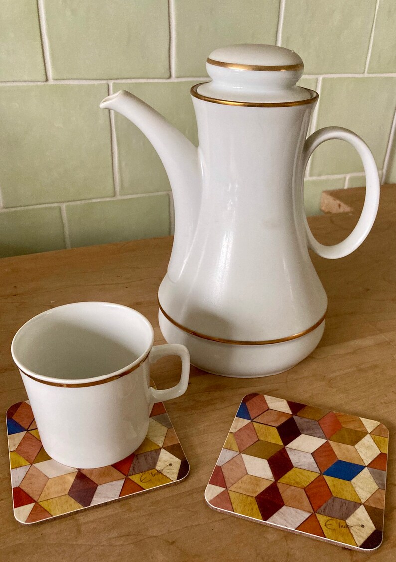 May include: A white ceramic coffee pot with a gold rim and lid, a matching cup, and two square coasters with a geometric pattern. The coasters feature a colorful mosaic design with various wood tones.