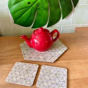 Puede incluir: Una tetera roja descansa sobre un posavasos hexagonal con un patrón geométrico, acompañada de dos posavasos cuadrados. Los posavasos presentan un diseño geométrico de color claro. Una gran hoja verde está en el fondo.