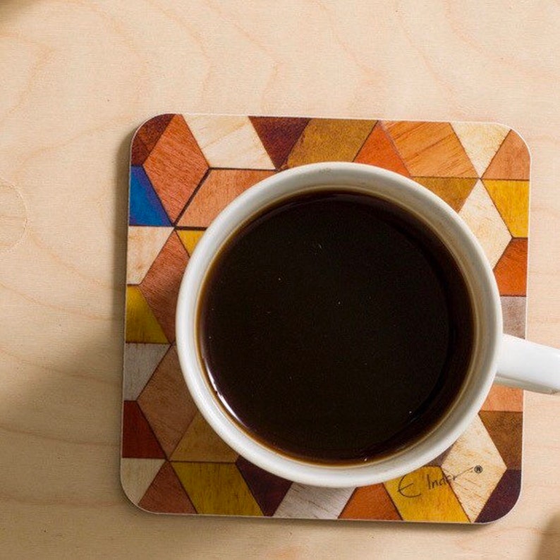 May include: A white ceramic mug filled with dark coffee sits on a square coaster. The coaster features a geometric pattern of triangles in shades of brown, orange, yellow, and blue. The mug is centered on the coaster, with a wooden surface in the background.