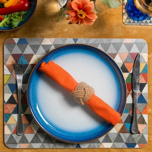 May include: A blue and white ceramic dinner plate with a light orange napkin tied with a brown rope. The plate is on a colourful geometric patterned placemat.