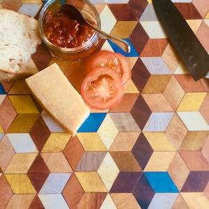 May include: A wooden chopping board with a geometric pattern in shades of brown, yellow, blue, and white. The board has a small dish of red sauce, a slice of bread, a piece of cheese, and tomato slices on it. A black knife is also on the board.