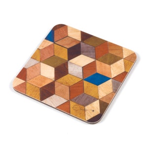May include: Square wooden coaster with a geometric cube pattern in shades of brown, tan, and blue. The coaster has rounded corners and a light-colored border. The design is made of various wood tones.