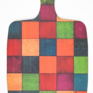 May include: A rectangular wooden cutting board with a rounded handle and a hole for hanging. The surface features a vibrant grid pattern of multicoloured squares in shades of red, orange, blue, green, and purple, with the signature E. Inder visible.
