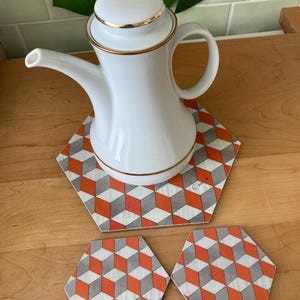 Op de afbeelding: Een witte porseleinen theepot met een gouden rand en deksel, rustend op een zeshoekige onderzetter met een geometrisch patroon in oranje, grijs en wit. Twee bijpassende onderzetters eronder.