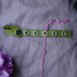 May include: A green leather needle holder with metal eyelets and a snap closure. A strand of pink yarn is threaded through one of the holes. A purple flower is in the lower left corner. The needle holder is on a gray linen surface.