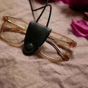 Puede incluir: Un par de gafas con montura transparente con un estuche de cuero negro para gafas sujeto por un cordón negro. Las gafas descansan sobre una tela arrugada de color beige. Pétalos de flores rosas esparcidos.