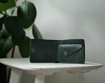 May include: Open, dark green leather wallet with a snap closure and multiple card slots. The wallet is displayed open, showcasing its interior design and stitching details. The wallet is sitting on a white surface.