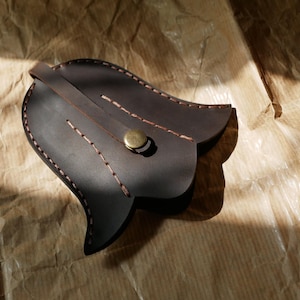 May include: A dark brown leather tulip-shaped pouch with a brass button closure. The edges are stitched with light brown thread. The pouch is lying on a crumpled brown paper surface.