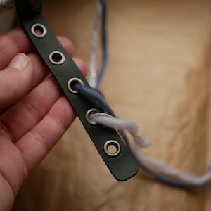Può includere: Un cinturino in pelle verde scuro con occhielli metallici e due fili di filo infilati negli occhielli. Il filo è bianco e blu.