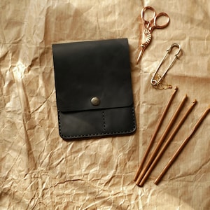 May include: A black leather pouch with a snap closure, next to a pair of decorative rose gold scissors, a safety pin, and several wooden crochet hooks. The items are arranged on a textured brown paper surface.