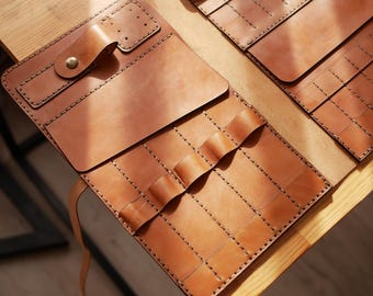 May include: A brown leather roll-up case with multiple slots for holding tools or other items. The case has a flap with a strap and a buckle closure.