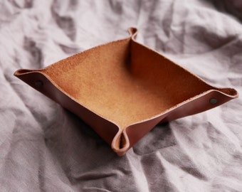 May include: A brown leather catchall tray with a square shape and folded edges. The tray is empty and has a soft, suede-like interior.