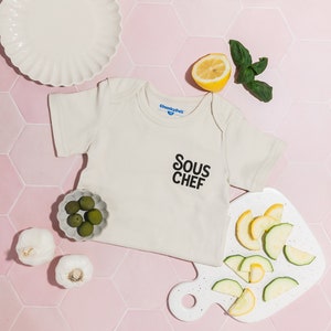 May include: A white baby onesie with the text "Sous Chef" printed in black. The onesie is on a pink tiled background with a plate, garlic, olives, a lemon, basil, and sliced courgette on a white chopping board.
