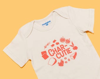 May include: A cream-colored short-sleeve baby bodysuit featuring a red graphic print with the text CHAR-CUTIE surrounded by illustrations of crackers, fruit, cheese, and fish. The inner neck label displays the brand name ChunkyDeli.