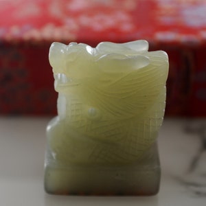 Op de afbeelding: Een klein, jadegroen drakenbeeldje met ingewikkelde details. De draak zit op een vierkante basis.