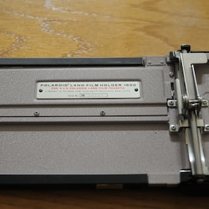 Może przedstawiać: Srebrny metalowy uchwyt do filmu Polaroid Land #500 dla 4 x 5 paczek filmu Polaroid Land. Uchwyt ma czarną ramkę i srebrny metalowy mechanizm przesuwny.