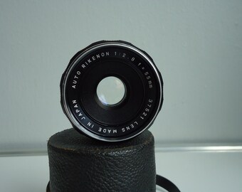 May include: A black camera lens with silver lettering that reads "AUTO RIKENON 1:2.8 f=55mm 37521 37521 LENS MADE IN JAPAN".