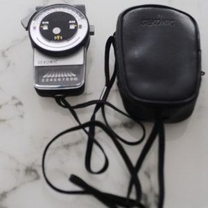 Puede incluir: Un fotómetro Sekonic plateado con una esfera negra y una funda de transporte de cuero negro. El fotómetro tiene una correa negra.