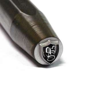 Könnte beinhalten: Nahaufnahme eines Metallstempels mit einem Schilddesign und den Buchstaben "QS" darin. Der Stempel ist silberfarben mit schwarzen Buchstaben.
