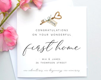 May include: A white card with a gold key with a heart-shaped bow on the handle. The key has a tag with the number 2024 on it. The card says "Congratulations on your wonderful first home" and "Mia & James, 26 Thompson Street". The card also says "new adventures, new beginnings, new memories".