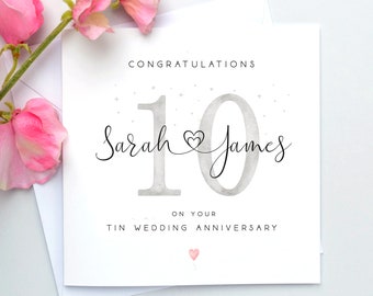 May include: A white greeting card with the text "CONGRATULATIONS" above the numbers "10" in silver. The names "Sarah & James" are written in cursive. The card is for a tin wedding anniversary, with a small pink heart at the bottom.