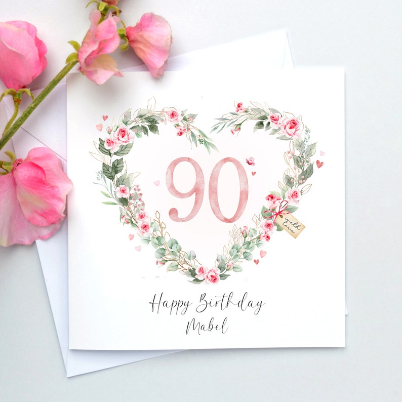 May include: A white card with a pink floral heart design. The number 90 is in the center of the heart. The text "Happy Birthday Mabel" is below the heart.