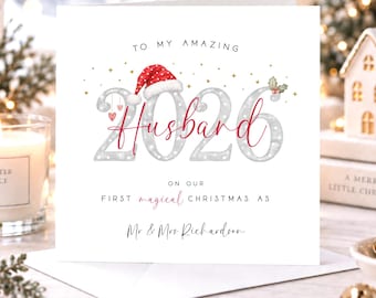 Peut inclure: Une carte de vœux carrée à fond blanc avec le texte TO MY AMAZING Husband ON OUR FIRST magical CHRISTMAS AS Mr & Mrs Richardson. Le nombre 2026 est imprimé en grande police grise avec un bonnet de Père Noël rouge et du houx.