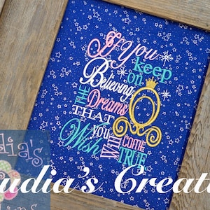 May include: Framed artwork with a blue background speckled with white stars. The artwork features the embroidered text "If you keep on believing the dreams that you wish will come true" in various colors, with a golden carriage design. The frame is made of weathered wood.