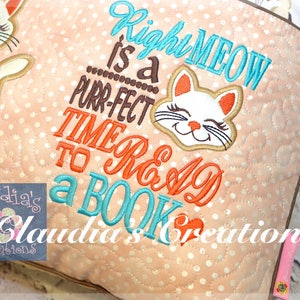 May include: A pink and white quilted pillow with a white cat face and the text "Right MEOW is a purr-fect TIME to READ a BOOK".