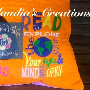 Könnte beinhalten: Ein orangefarbenes, gestepptes Kissen mit dem Text "READ to EXPLORE the world with your eyes & MIND wide OPEN." Ein kleineres Patch zeigt den Text "Claudia's Creations" und ein Blumenmuster.