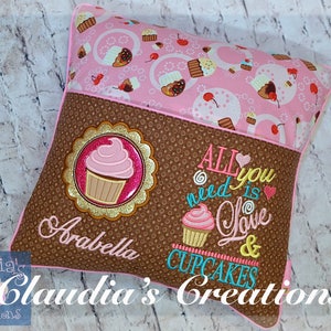 May include: A decorative pillow with a pink and brown cupcake theme. The pillow features a pink cupcake design, the words "All you need is Love & Cupcakes", and the name "Arabella". The pillow is trimmed in pink.