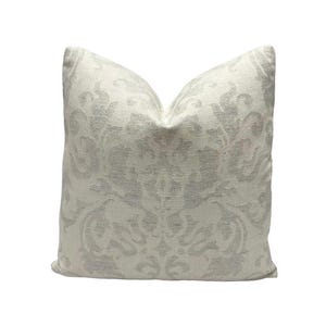 May include: A square decorative pillow with a light cream background and a subtle, repeating gray floral pattern. The pillow has a soft, textured appearance and is designed for home decor.