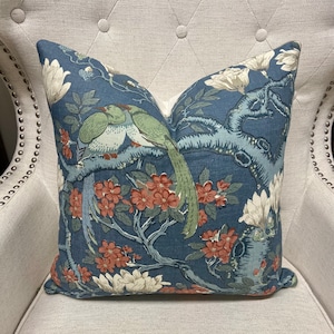 May include: Decorative throw pillow with a blue background featuring a floral and bird design. The pillow has a pattern of green birds, red flowers, and white blossoms. The pillow is placed on a light-colored upholstered chair.