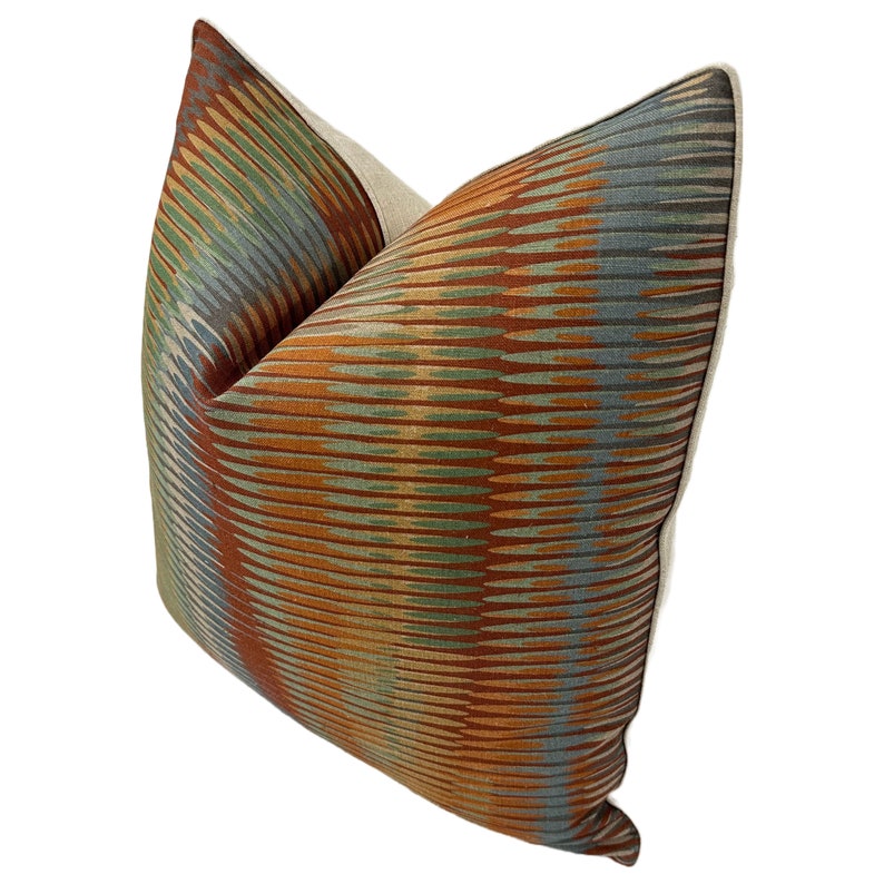 Puede incluir: Una almohada decorativa con un patr&oacute;n geom&eacute;trico en tonos naranja, marr&oacute;n, verde y azul. La almohada tiene un borde de tela de lino beige.
