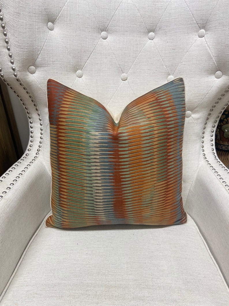 Puede incluir: Una almohada decorativa con un patr&oacute;n abstracto colorido en tonos naranja, verde, azul y beige. La almohada est&aacute; sentada en una silla blanca.