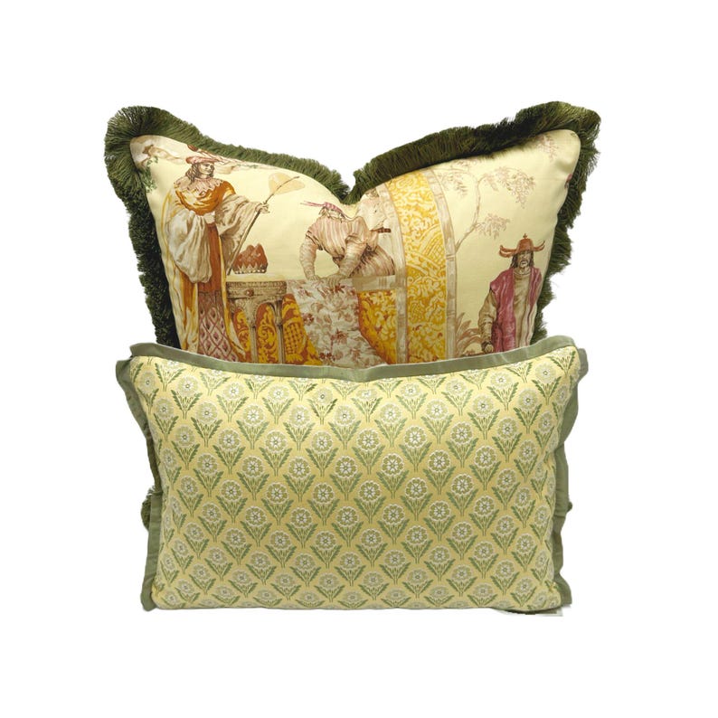 May include: Two decorative throw pillows with a green fringe. The top pillow has a yellow background with a colorful print of people in a garden setting. The bottom pillow has a yellow background with a green geometric pattern.