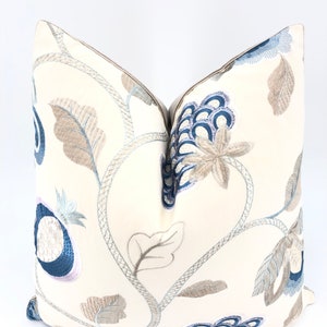 Puede incluir: Una almohada blanca con diseños florales bordados en azul y marrón. La almohada tiene un borde de color crema.