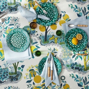 May include: A table setting with a botanical theme. The white tablecloth features a pattern of yellow lemons, blue leaves, and butterflies. Green plates, blue napkins, and bamboo-handled silverware are arranged for dining. Two lemons are on one plate.