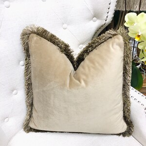 May include: A beige velvet pillow with a brown fringe border. The pillow is sitting on a white tufted chair.