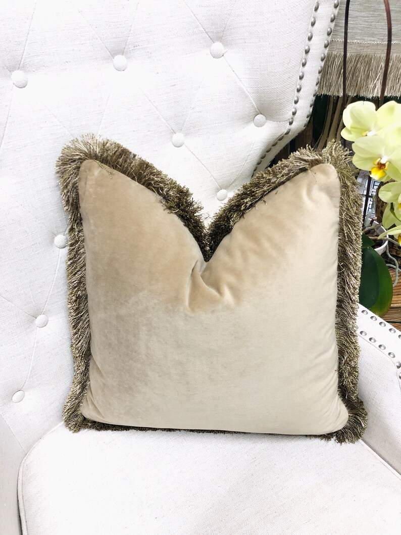 May include: A beige velvet pillow with a brown fringe border. The pillow is sitting on a white tufted chair.