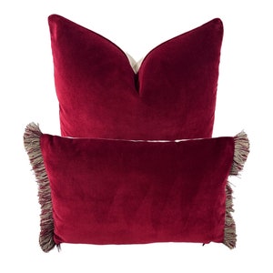 May include: Two red velvet throw pillows with fringe trim. The top pillow is a standard square shape, while the bottom pillow is rectangular.