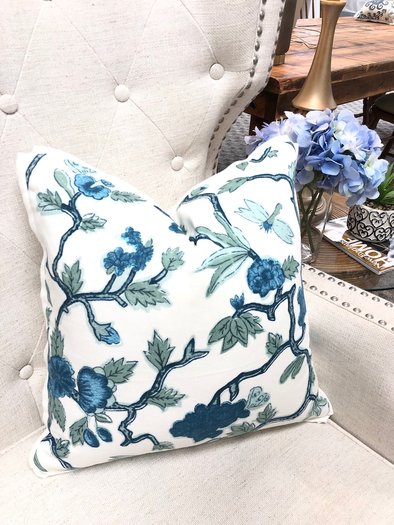 May include: A white throw pillow with a blue and green floral pattern. The pillow is sitting on a white armchair with tufted buttons.